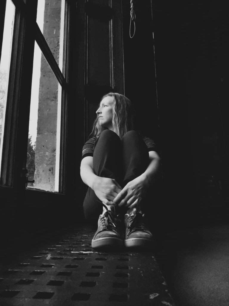 Chrissie sitting by window, looking out. Arms around knees