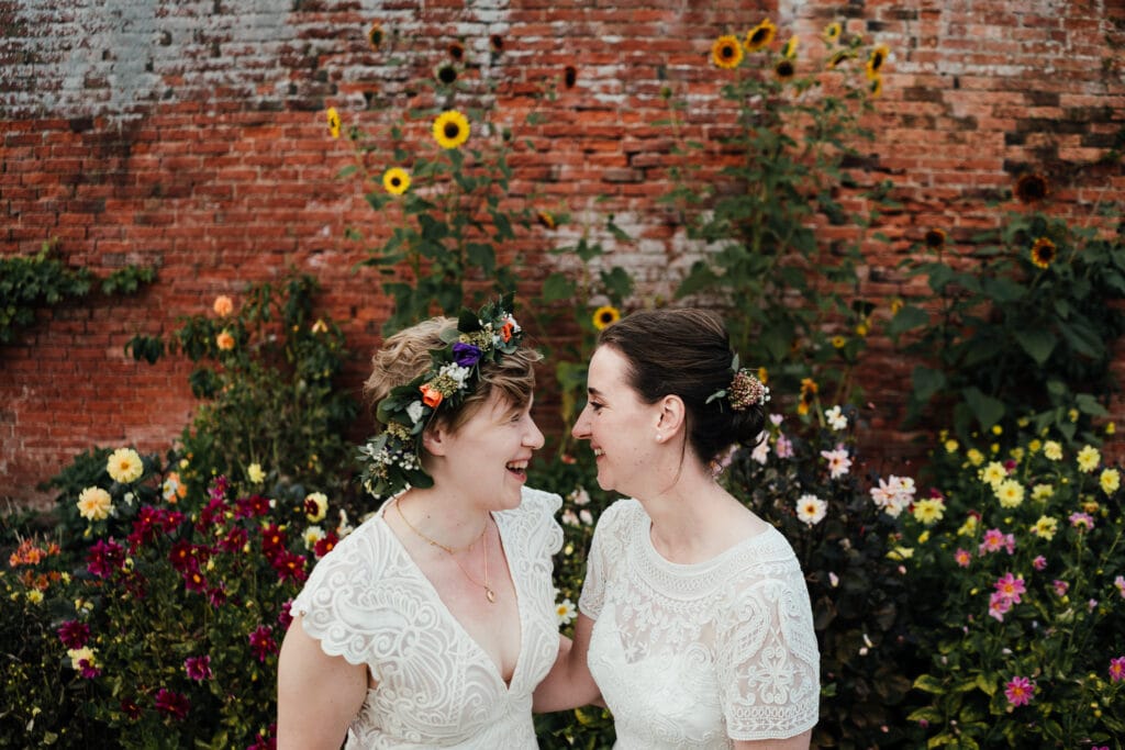 Wedding photography at Thorpe Garden. Brides laughing together in front of walled garden, the wall adorned with pretty flowers.
