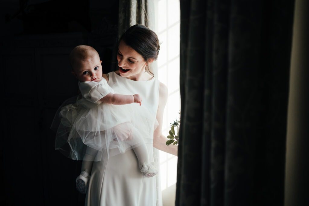Bride holding baby daughter in window light.