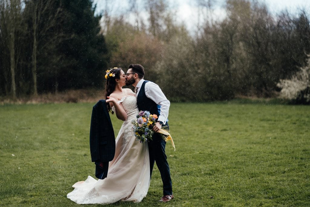 Bride and groom kiss in the rain. Groom holds bride's bouquet, bride holds groom's jacket. In the woodlands of the Hidden Hive