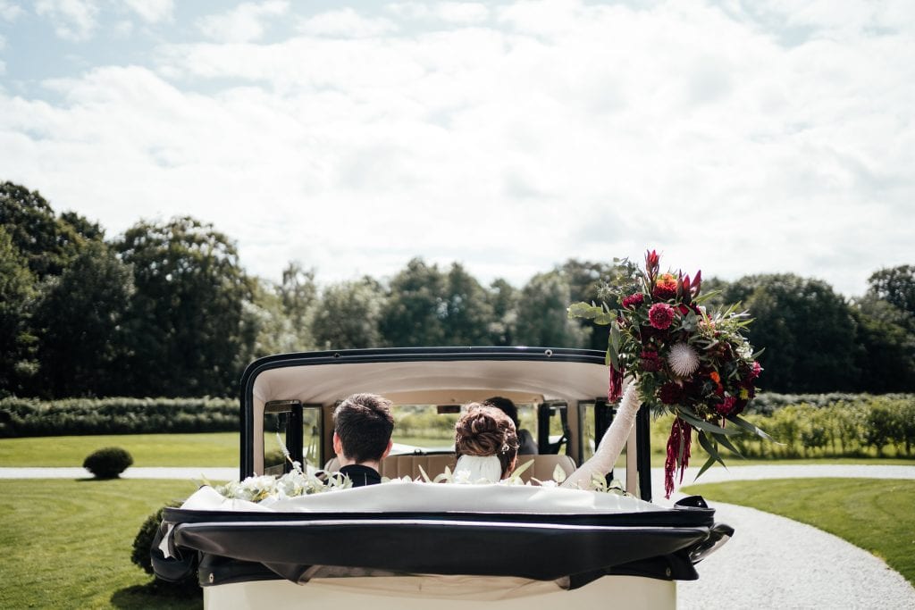 Bride and groom in back of vintage car as it drives off. Rood down, bride raising her bouquet