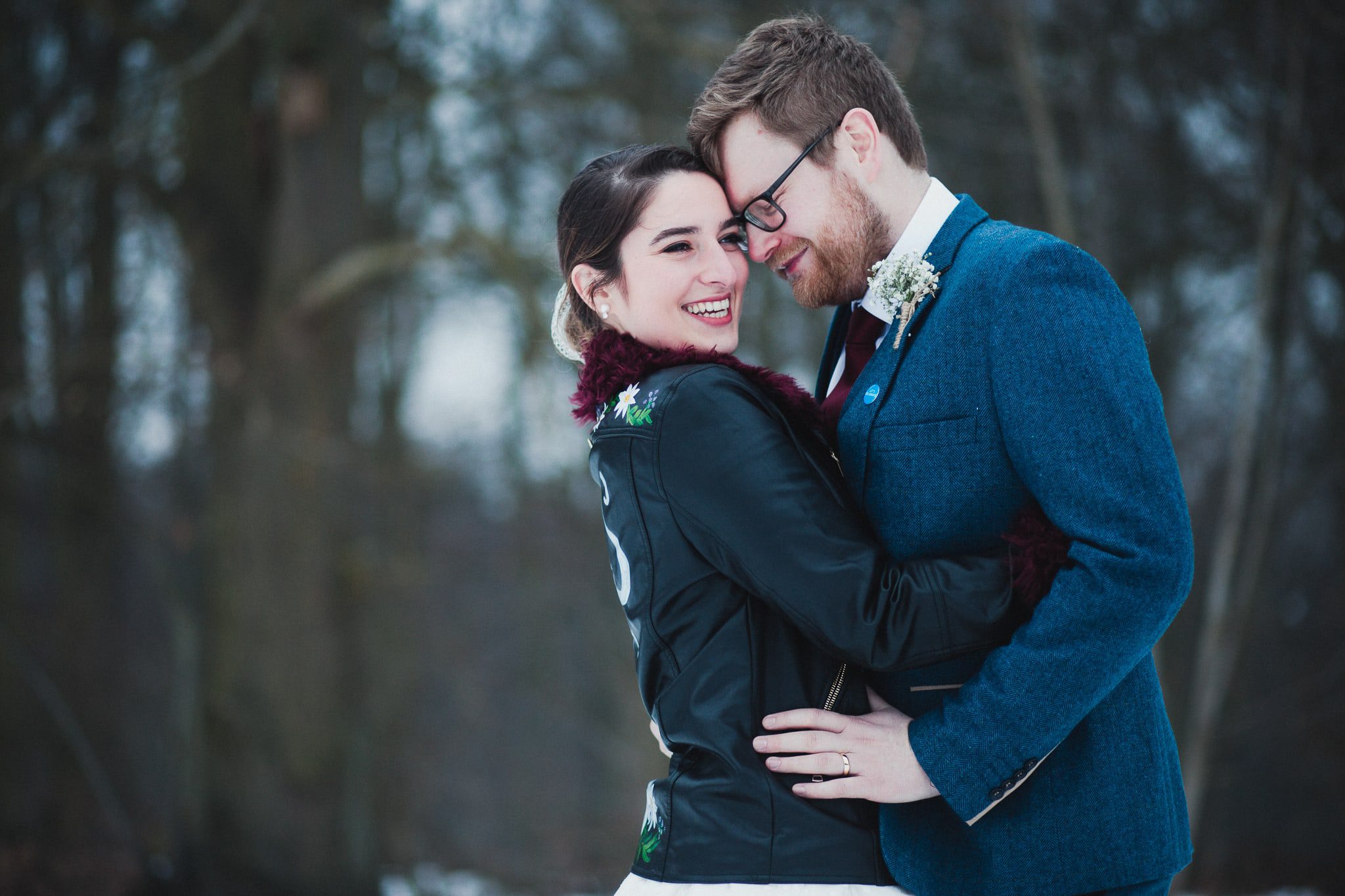 Bride and groom mid shot portrait. groom's head resting against bride's. Bride in leather jacket. Against a snowy backdrop and woods at Hazel Gap Barn blurred behind them.