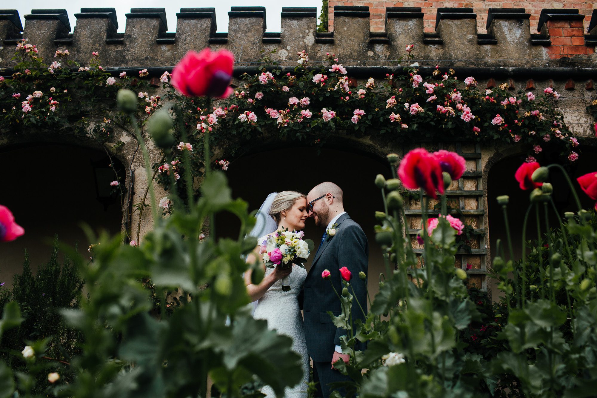 Bride and groom in walled garden at Holme Pierrepont Hall, surrounded by red poppies and pink roses in the courtyard