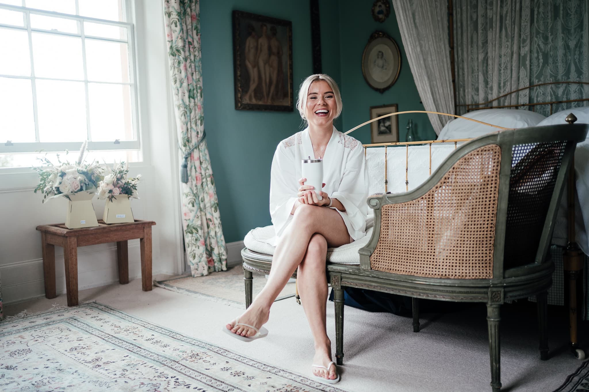 Bride sitting on the chaise long at Langar Hall in her dressing gown, laughing