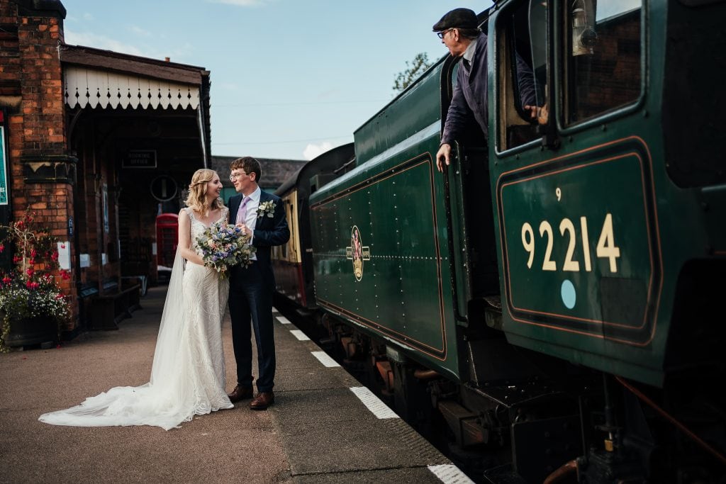 Bride and groom by railway with steam train rolling past. Conductor looking out of window at them.