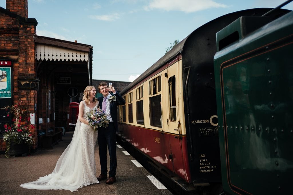 Bride and groom by railway. Groom raises champagne glass to train driver as they pass.