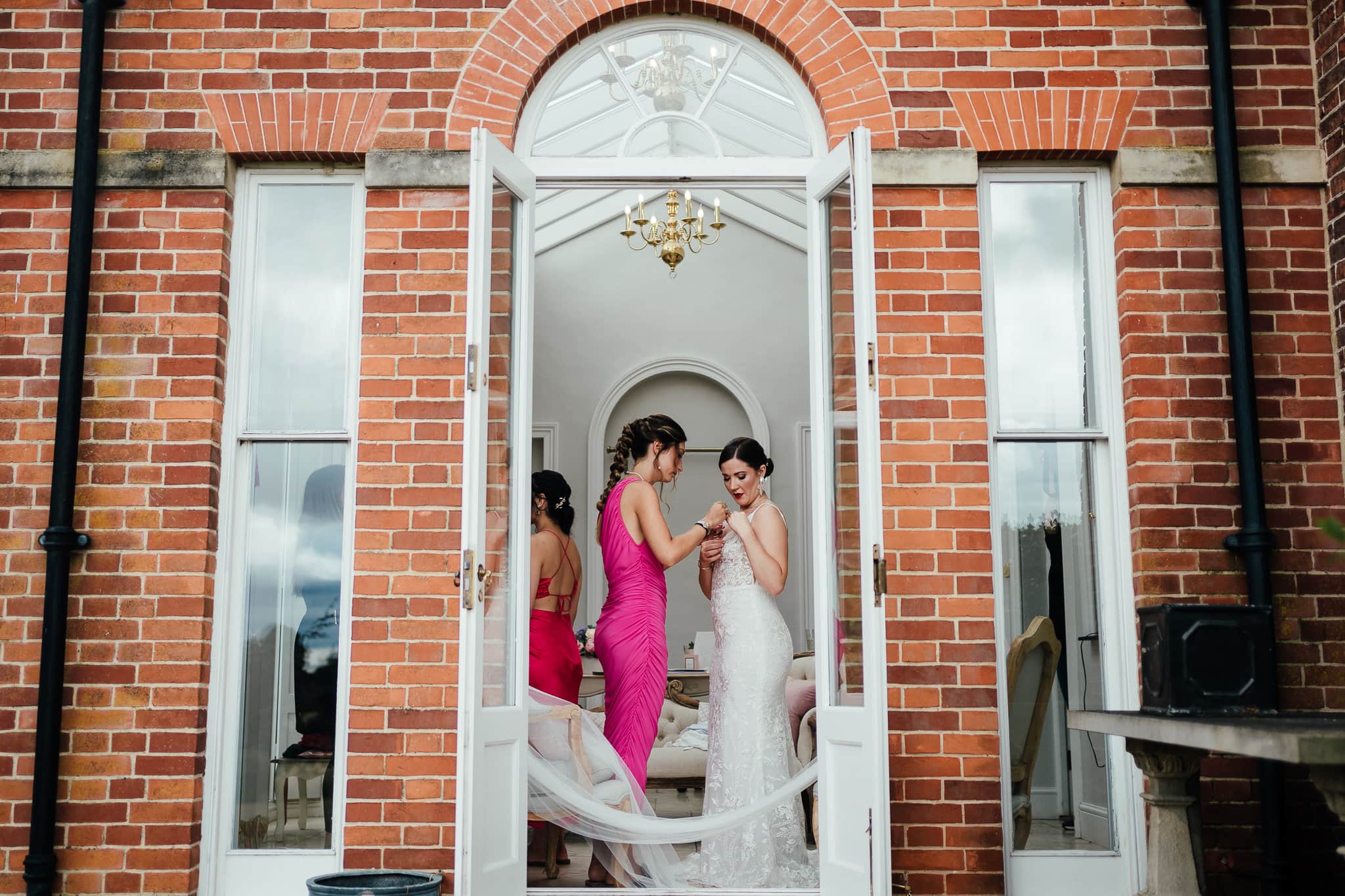 In the French doors of the bridal preparations room, a bridesmaid dressed in pink helps the bride with her dress.