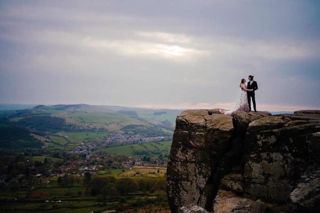 Bride and groom stand on top of the big rock at Curbar Edge, with the rolling Peak District landscape falling behind them.