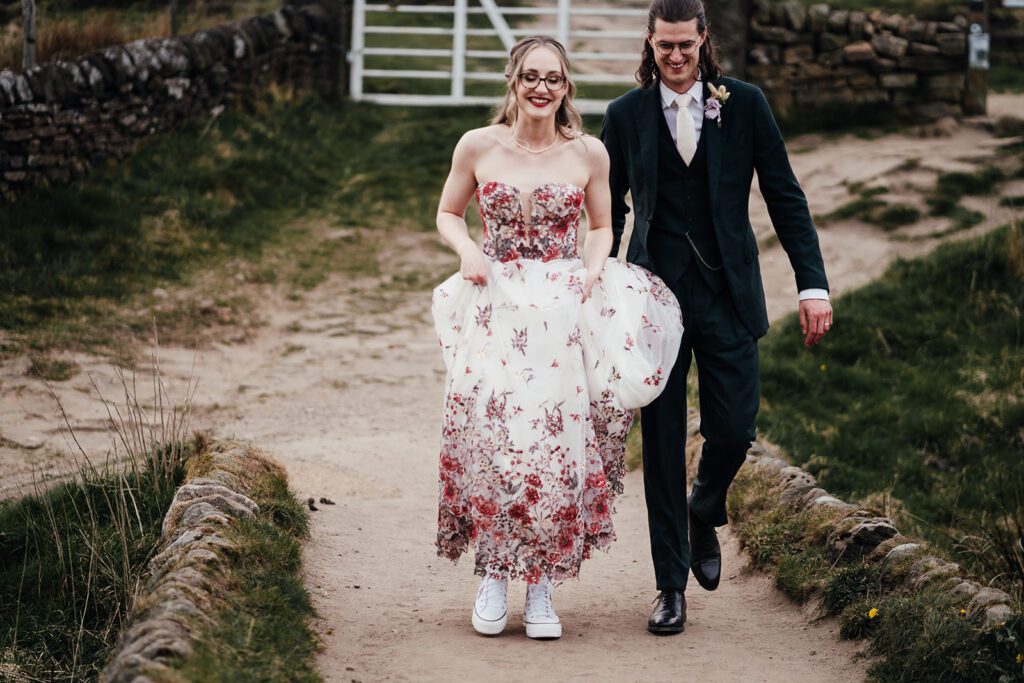 Bride and groom walking across Curbar Edge in the Peak District. Bride holds wedding dress above her ankles, revealing her white converse under her dress.