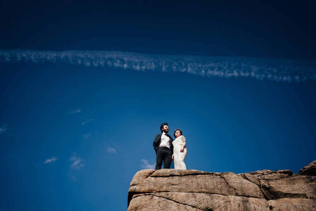 Bride and groom stand on a rock with blue sky behind them