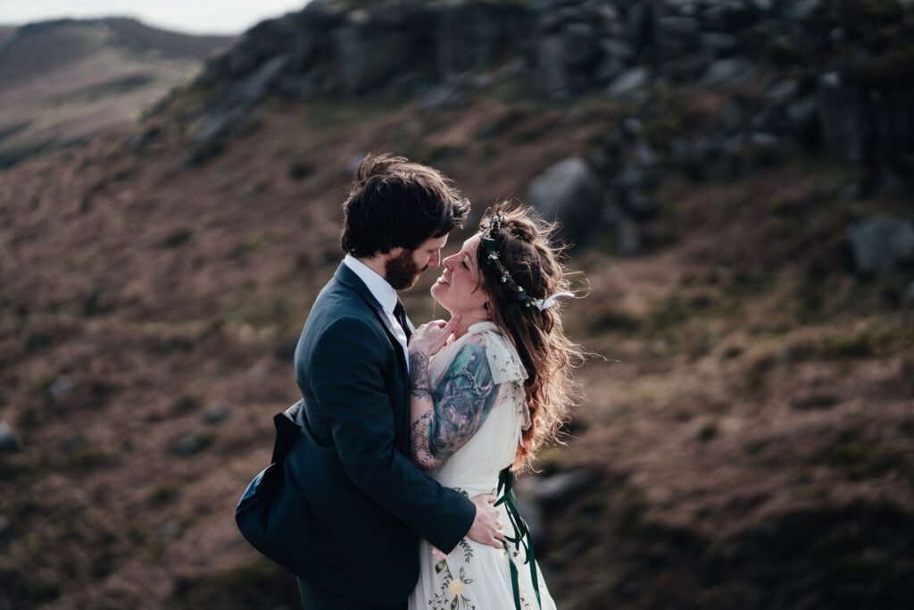 Bride and groom embrace on a rock in Burbage Peak District, the rolling hills behind them