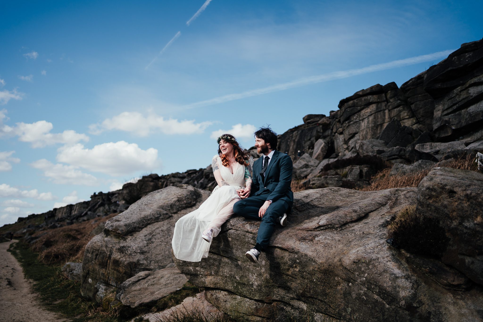 ride and groom sit on a rock against the rugged landscape of the Peak District. Bride is laughing as she looks out to the distance. Her flowing dress draped over the rocks. She holds onto groom's hand. Grey rocks of the peak disctric and a blue sky are behind them.