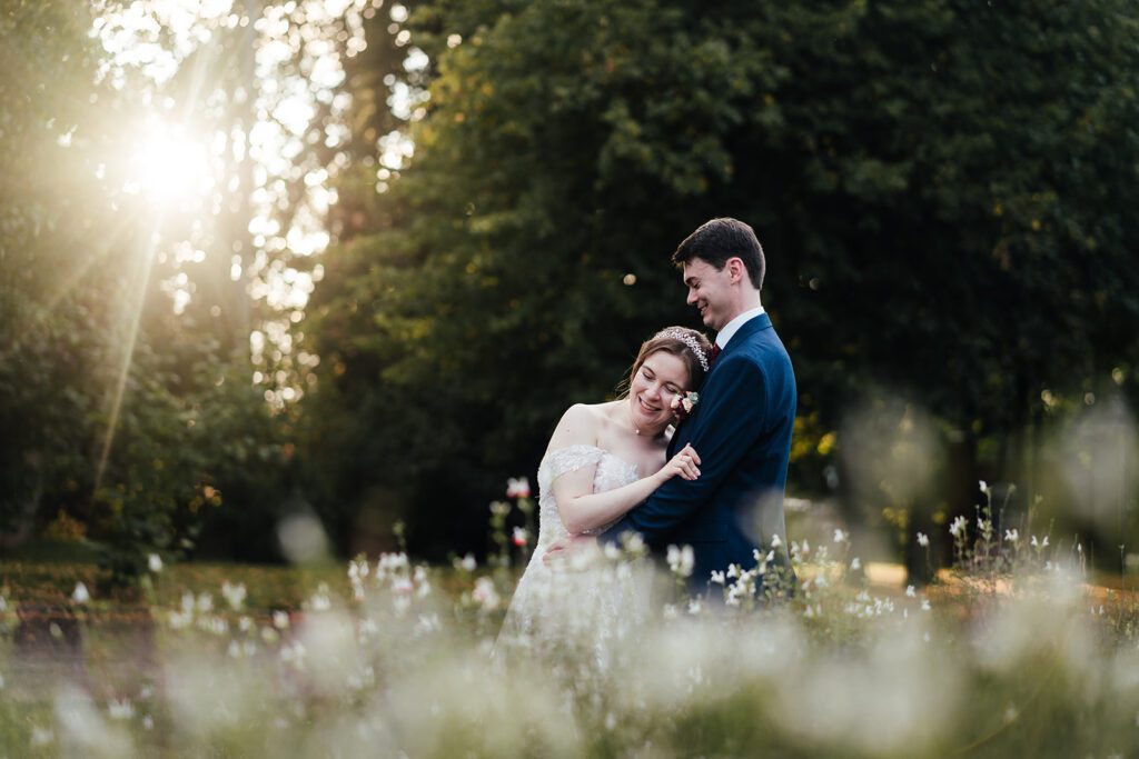 Colwick Hall wedding photographer - bride and groom standing in the flowers outside Colwick Hall with the sun shining behind them. Bride's head is resting on groom's chest. Groom is smiling at her.