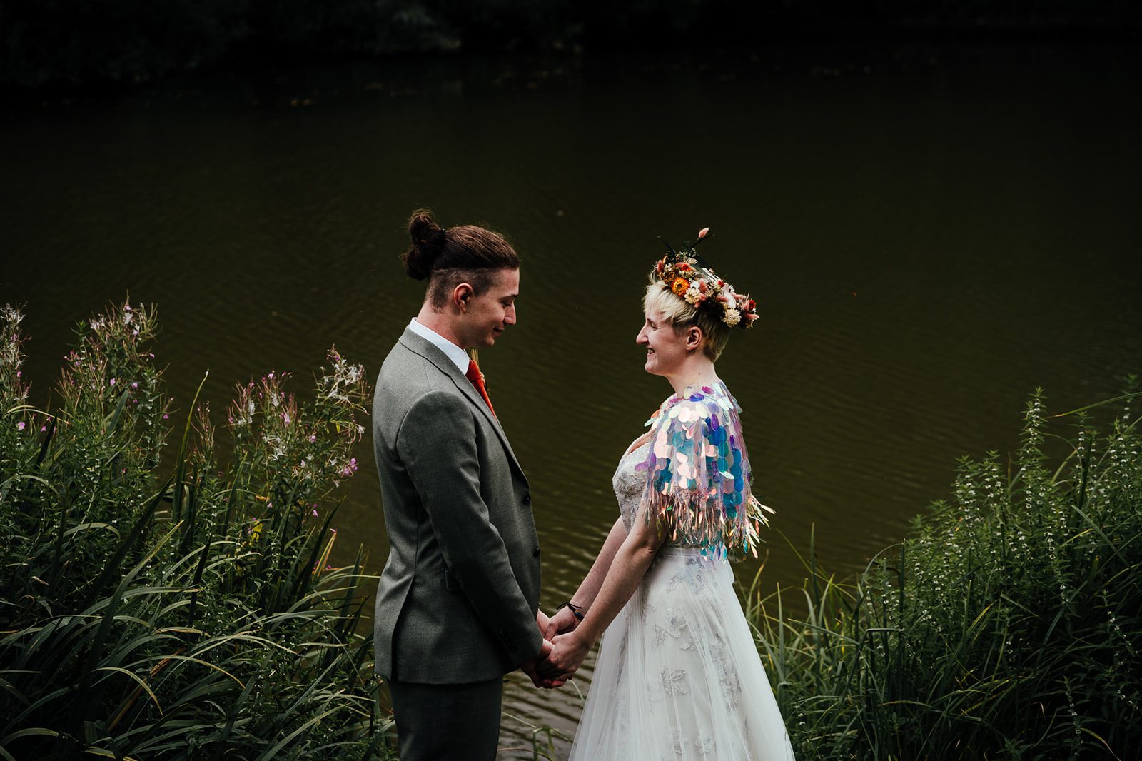 Bride and groom at The Hidden Hives Tipis. In front of the lake, looking at each other. Bride has a flower crown of dried flowers and wears a sequinned bolero over her wedding dress. Groom is in an olive green suit.