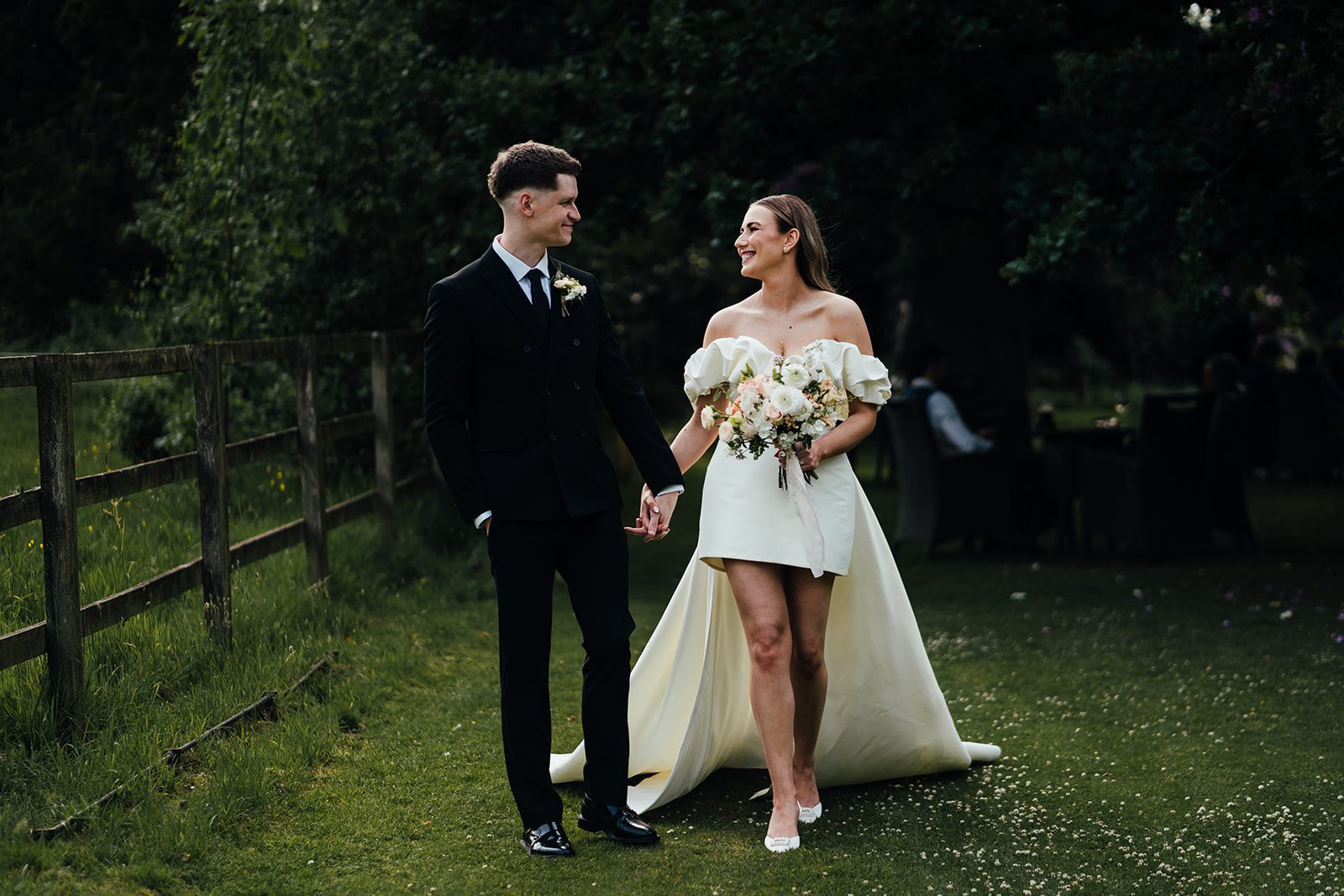 Bride and groom at Woodborough Hall wedding venue, walking in the grounds hand in hand and looking at each other. Bride wears a short wedding dress with a long train, groom wears a black suit and black tie.