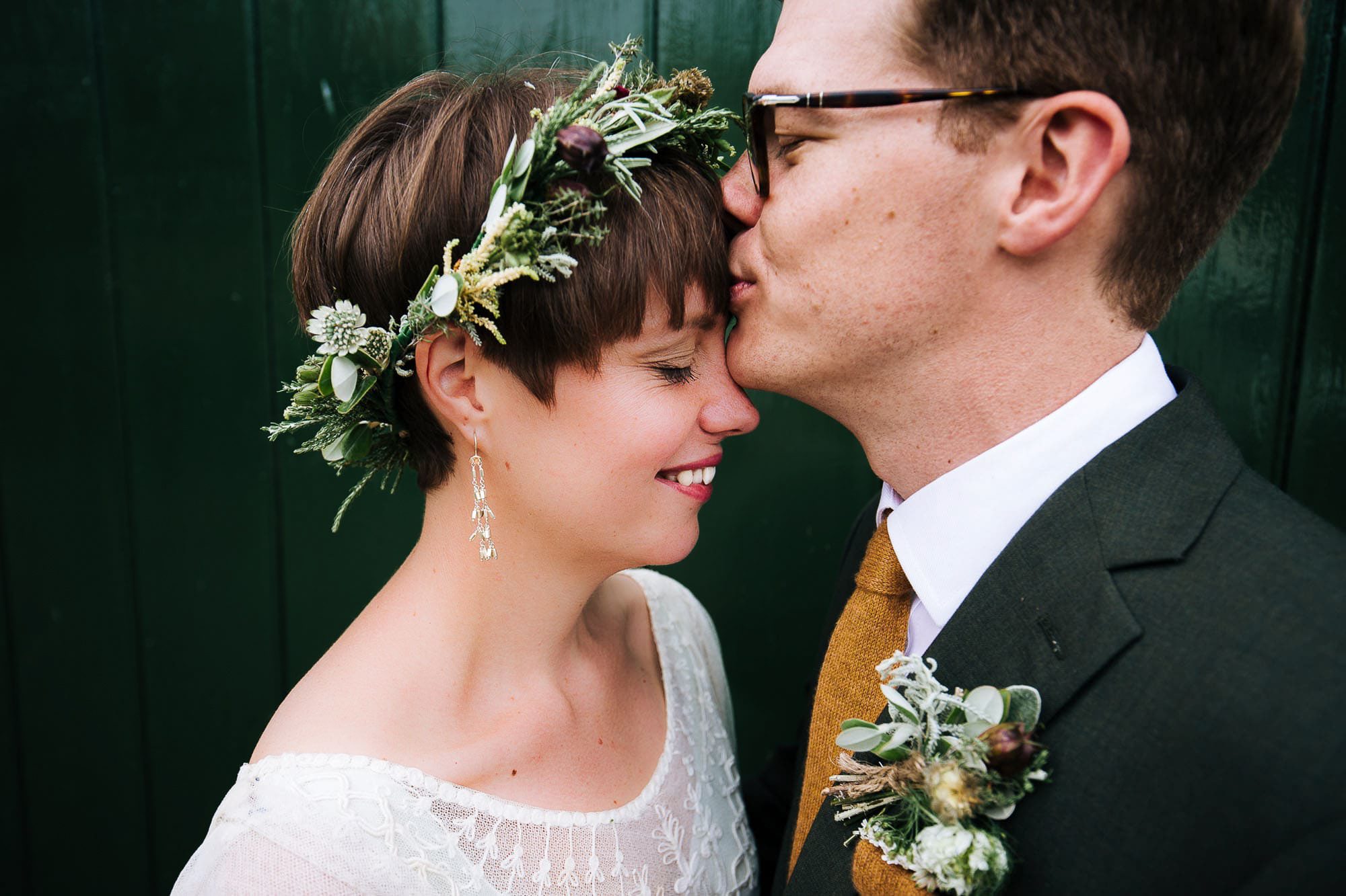 Close up of bride and groom. Bride with short hair and flowersr crown, eyes closed and smiling, groom kissing her head
