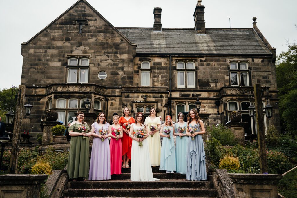 Bride and Bridesmaids in pastel coloured dresses standing on the steps outside Hargate Hall.